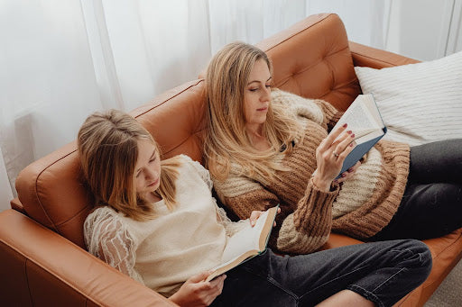 Two women, possibly a mother and daughter, comfortably reading books while reclining together on a brown leather couch, enjoying a quiet moment.