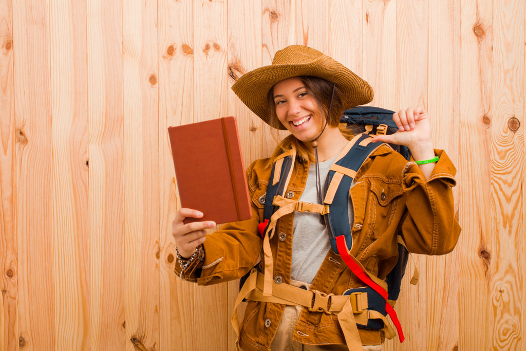 Adventurous young woman in hat smiling with book against wooden backdrop.