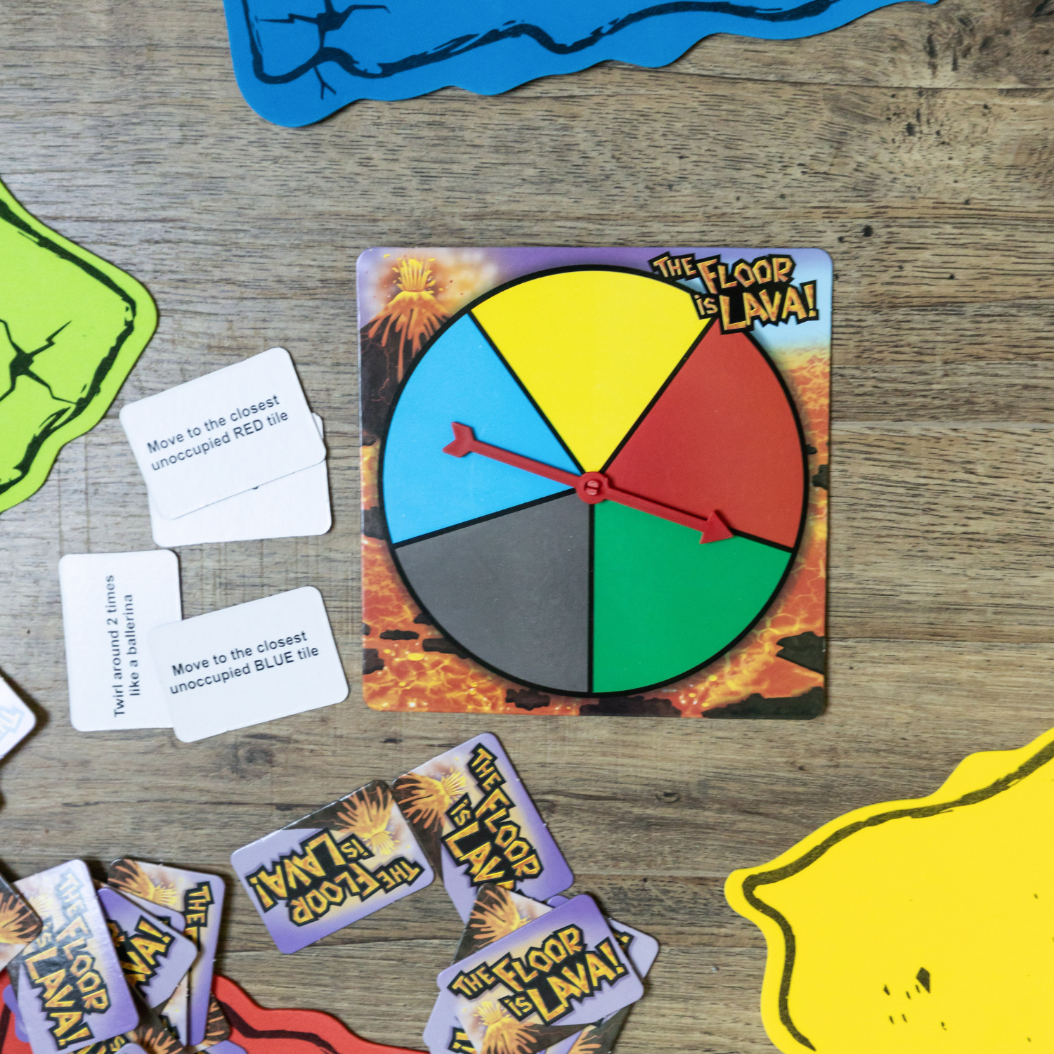 A multi-colored wheel spinner is in the center of the image next to a pile of cards. The spinner has an image of a volcano in the background and says The Floor is Lava.