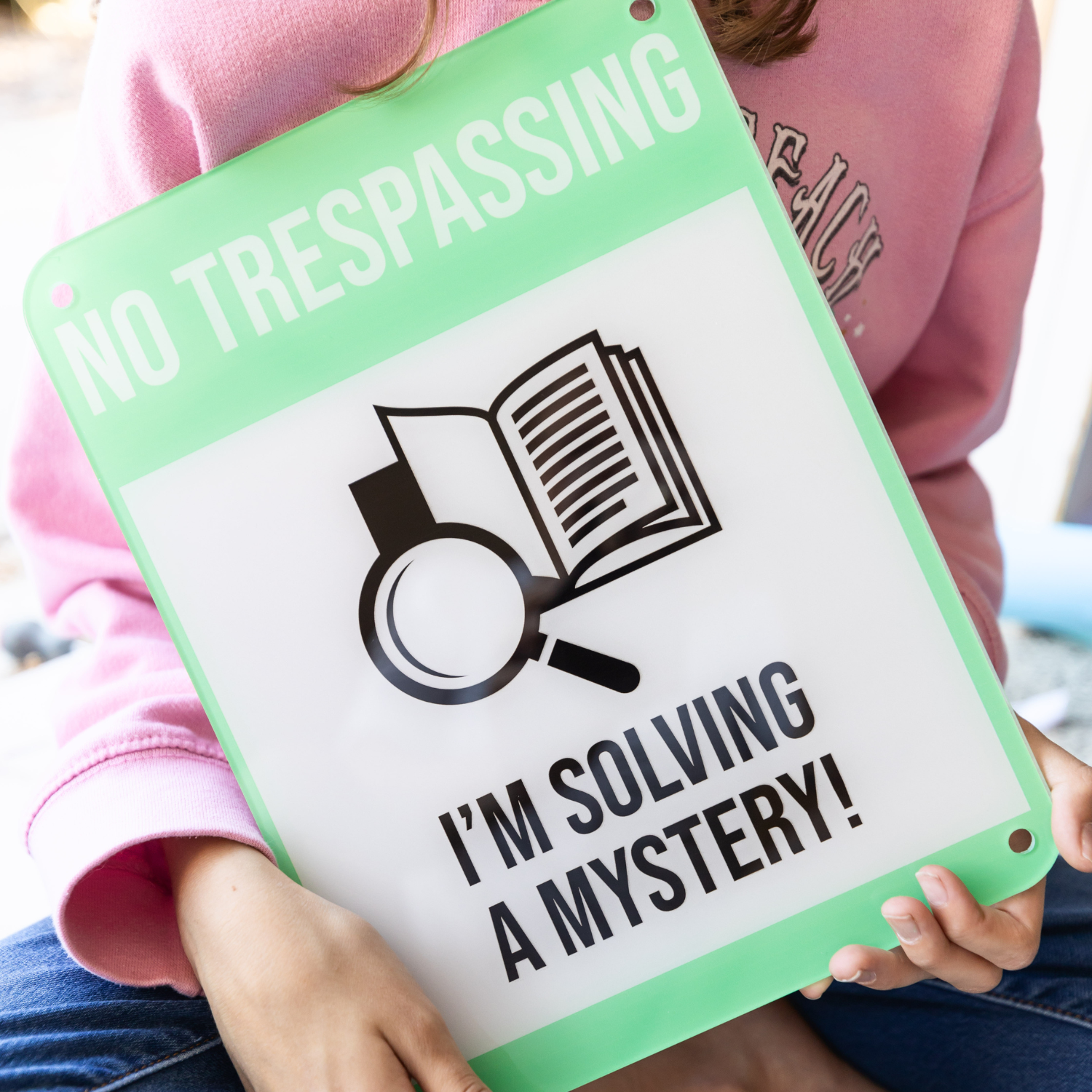 A girl wearing a pink sweatshirt holds a green and white sign that says No Trespassing I'm Solving a Mystery! with an image of a magnifying glass in front of a book at the center of the sign.