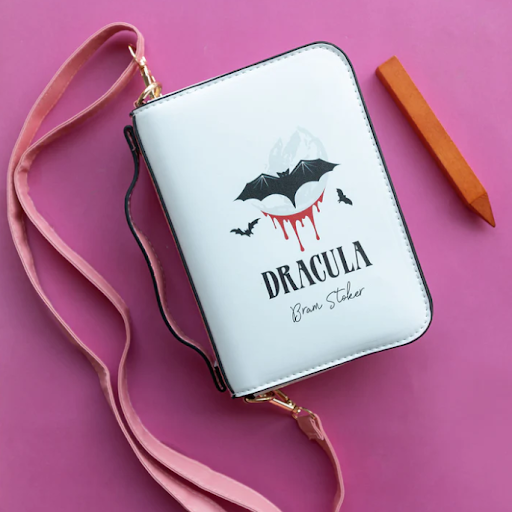 A stylish purse designed to resemble the cover of 'Dracula' by Bram Stoker rests on a vibrant pink surface. The purse is white with a black bat design, red droplets, and the title 'Dracula' in black letters. A blush pink strap is attached to the purse, and there's a brown pencil lying beside it.