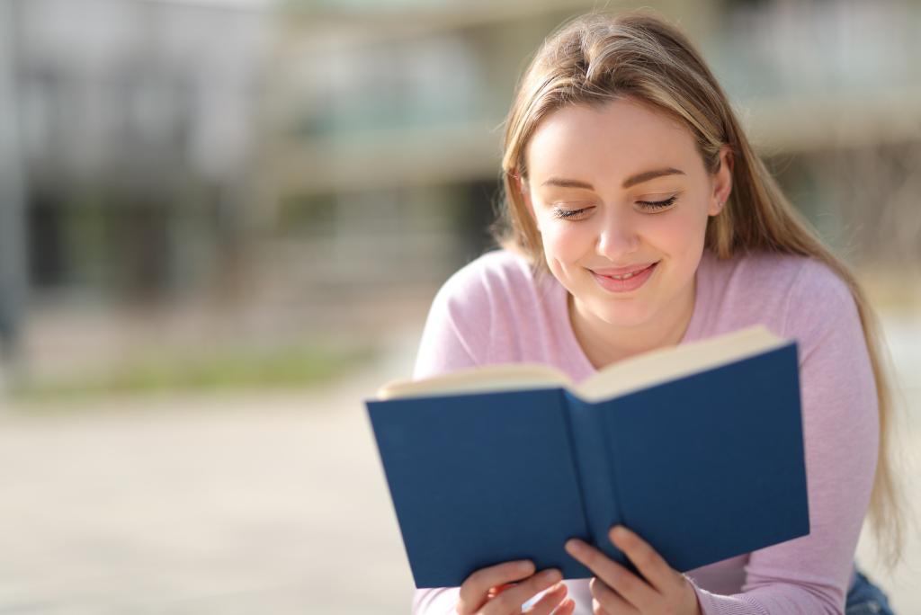 A young woman smiling while reading a book outside.