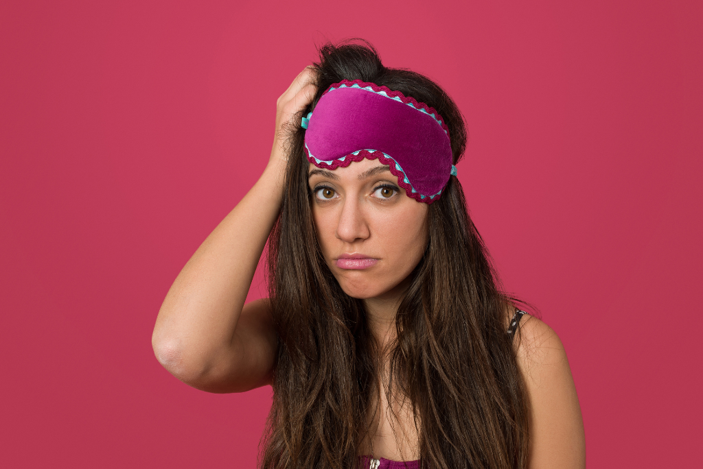 Woman with a sleep mask on her forehead looks tired against a pink background.