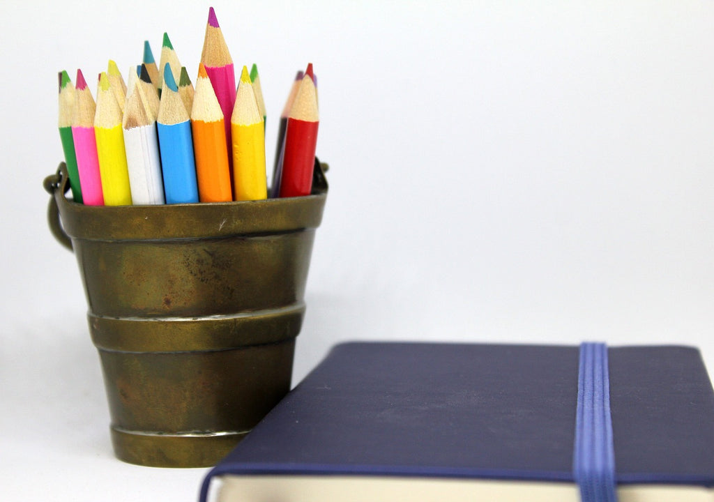 Colored pencils in a rustic brass bucket beside a closed navy blue notebook with an elastic band, set against a white background, suggesting creative writing or artistic endeavors.