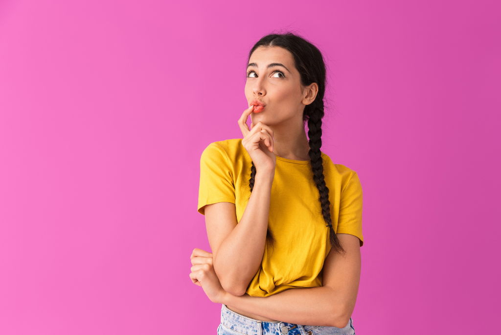 Thoughtful woman with braid against a pink background, finger on lip.