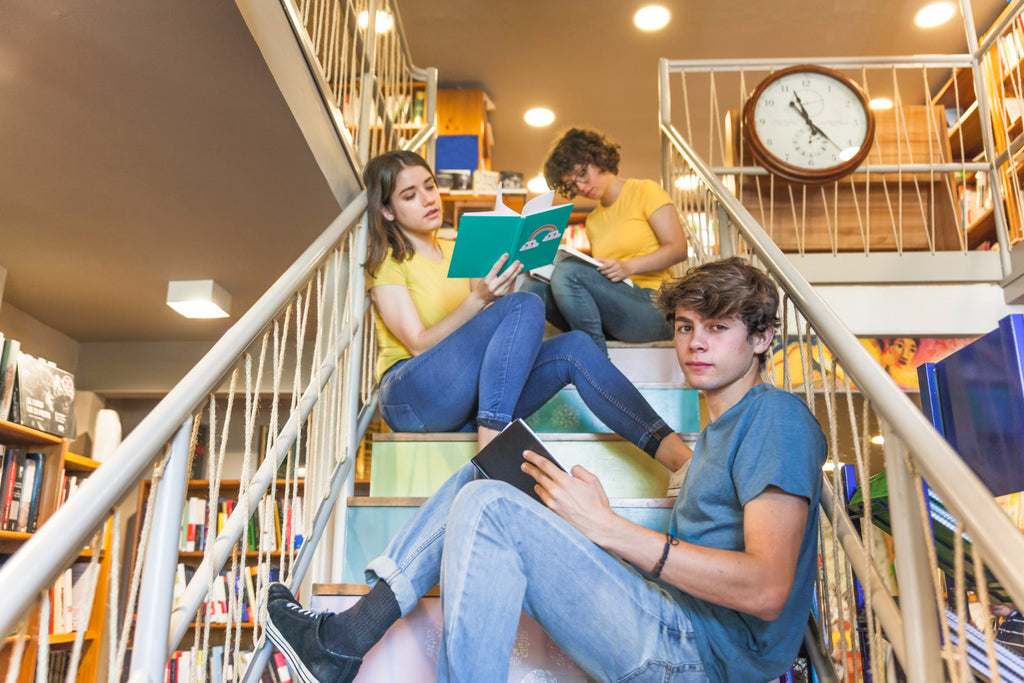 Three young adults reading books on library stairs, with a clock and shelves in the background.