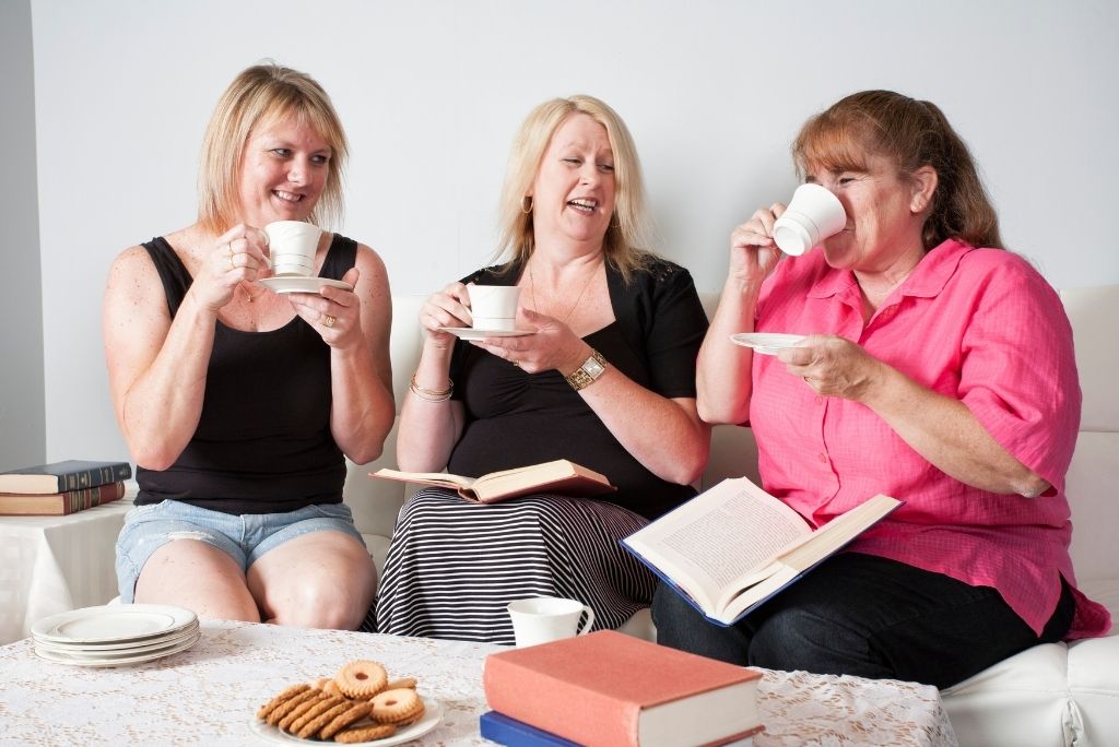 Three women laugh and share stories over tea and books, enjoying a casual book club meeting.