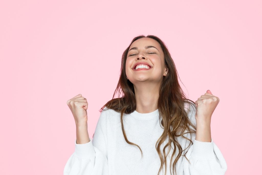 A woman in a white sweater closes her eyes and smiles with joy, her fists raised in a celebratory gesture against a pink background.