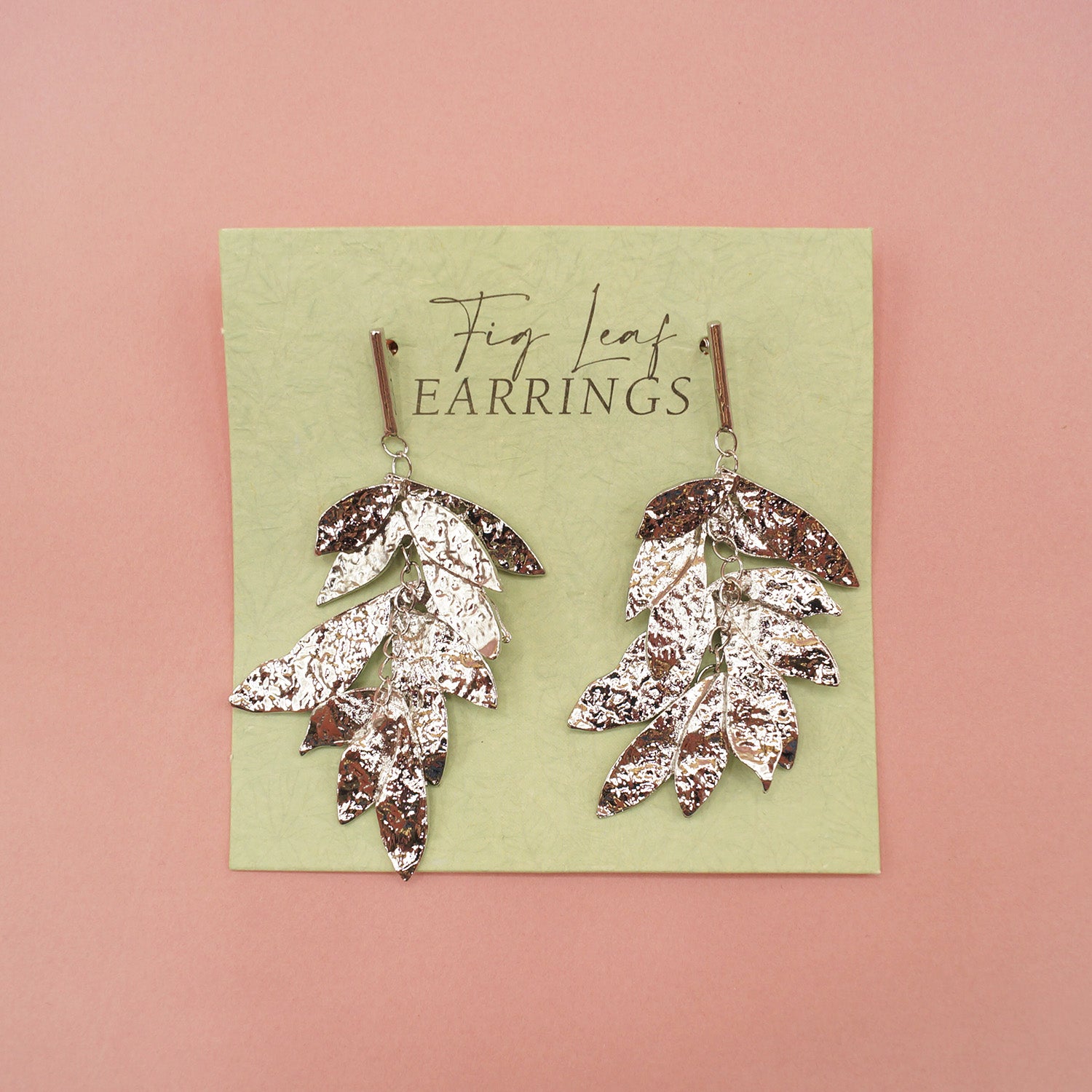 a pair of silver fig leaf earrings sits on a pink background