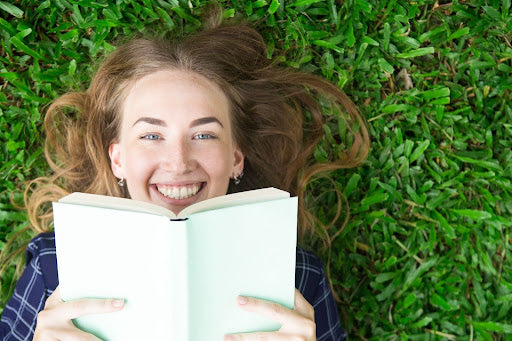 A joyful young woman with her hair spread out lies on lush green grass, holding an open book up to her face and laughing. The book obscures the lower half of her face, highlighting her twinkling eyes and the expression of delight.