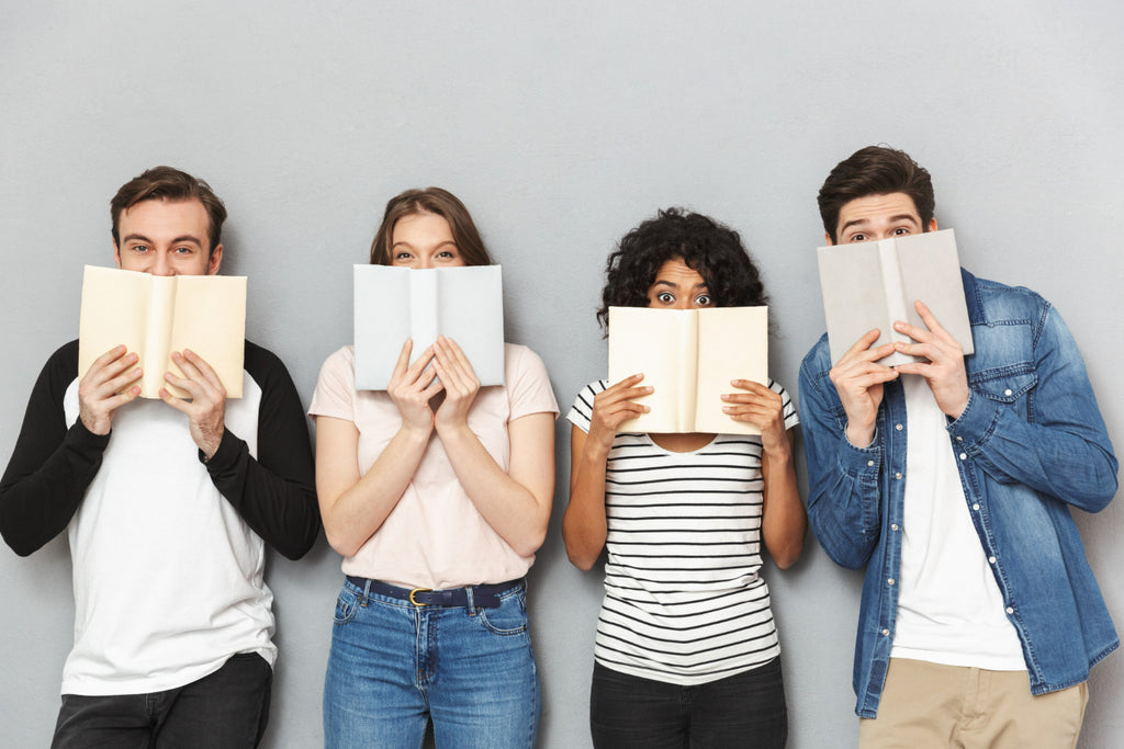 Four young adults stand against a grey background, each with a book covering half of their face, showcasing a mix of playful and intrigued expressions, symbolizing the diverse appeal of reading.