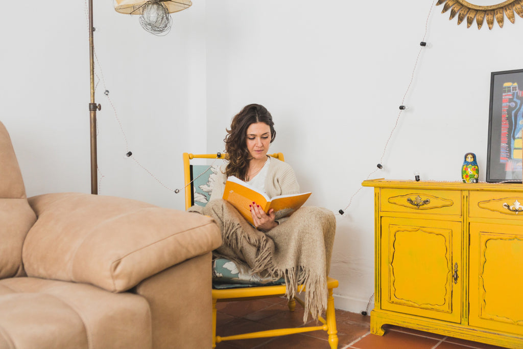 Woman reading a book on a yellow chair with a beige throw blanket.