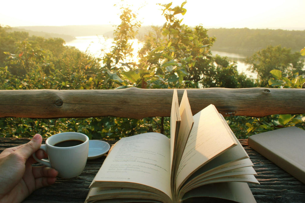 A serene reading scene with a hand holding a cup of coffee, an open book, and a closed notebook on a rustic wooden table with a view of a tranquil river and lush trees in the warm light of sunset.