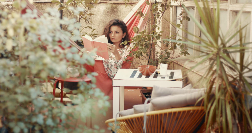 A woman reads a book in a cozy hammock on a plant-filled balcony.