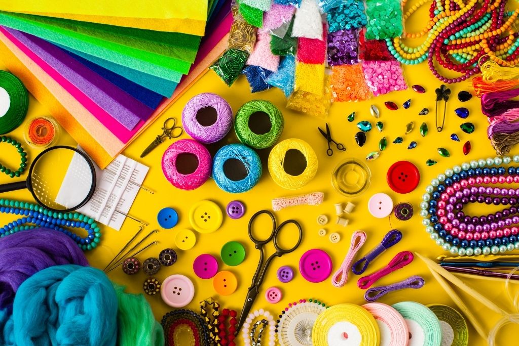 Colorful crafting materials spread out, including beads, buttons, yarn, ribbons, and paper on a bright yellow background.