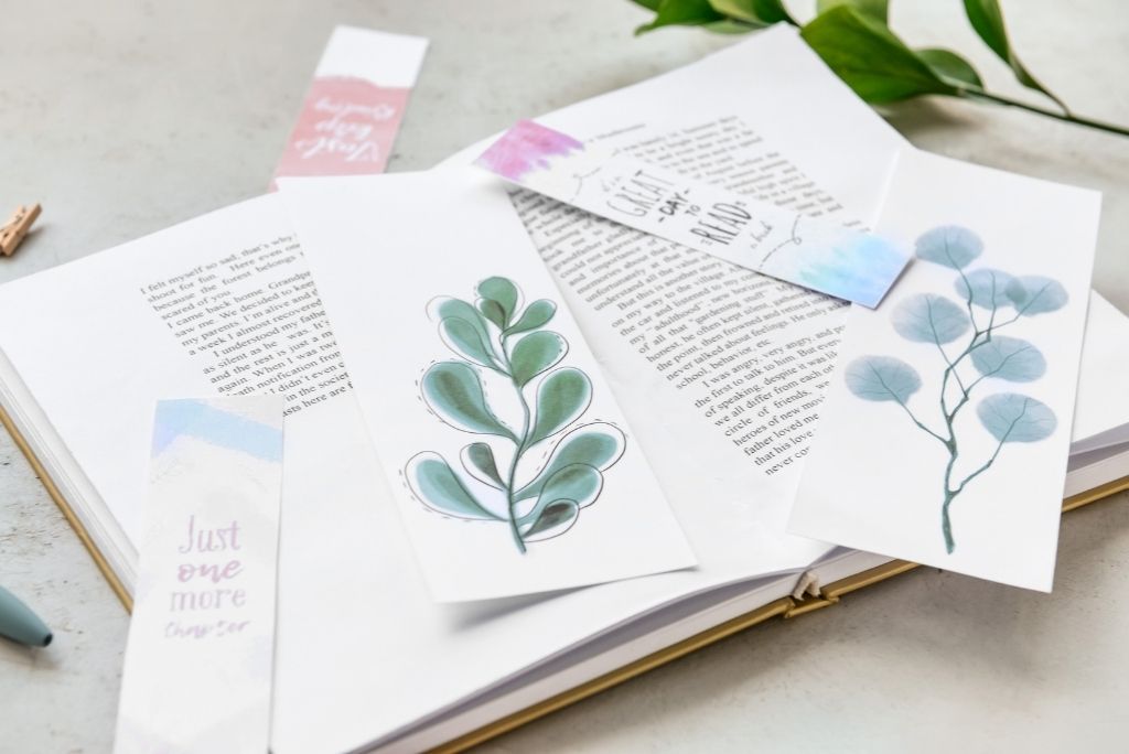 Handcrafted bookmarks with delicate watercolor illustrations and quotes lying on an open book beside a green plant.