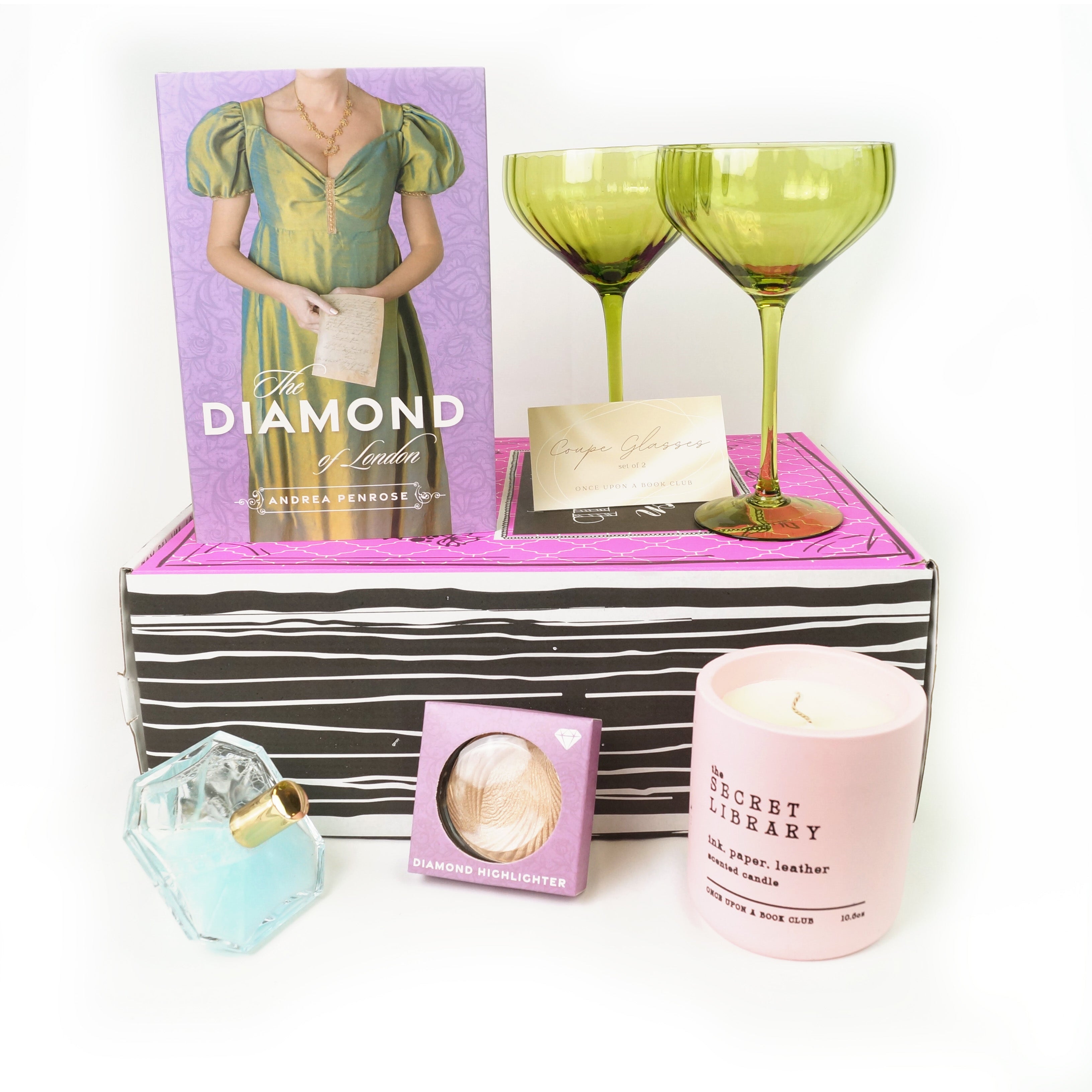 A hardcover special edition of The Diamond of London stands on a pink OUABC box next to a pair of green coupe glasses. In front of the box are a diamond-shaped glass container with light blue liquid, highlighter, and a pink candle labeled The Secret Library.
