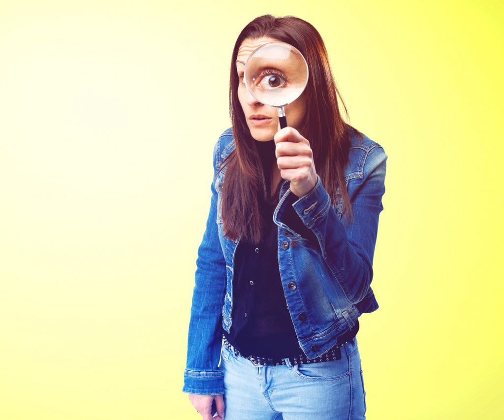 Woman in a denim jacket holding a magnifying glass up to her eye, with a yellow background.