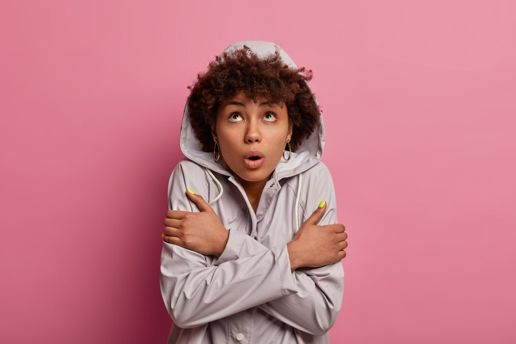 Surprised young woman with curly hair wearing a hooded jacket, pointing in two different directions against a pink background.