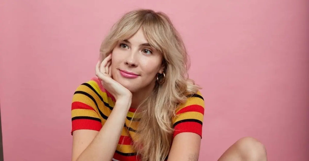 Smiling woman with blonde hair, resting her chin on her hand, wearing a colorful striped shirt, sitting against a pink background.
