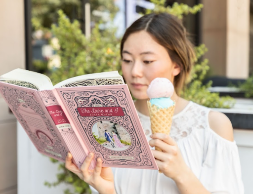 A young woman enjoys reading "The Duke and I" while savoring a two-scoop ice cream cone, embodying the pleasure of indulging in a good book and a sweet treat outdoors.
