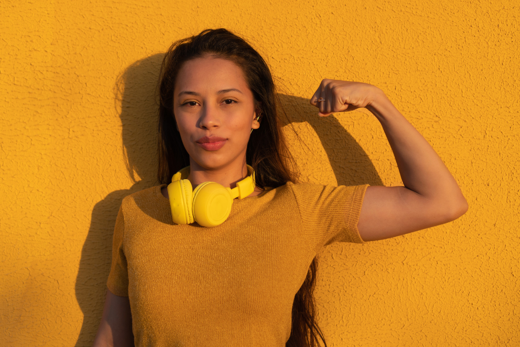 Confident young woman flexing her arm muscles, wearing yellow headphones and a mustard top, against a vibrant yellow wall