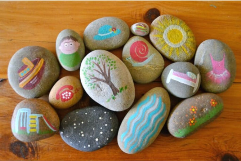 Painted stones with colorful illustrations of various objects and characters laid out on a wooden table.