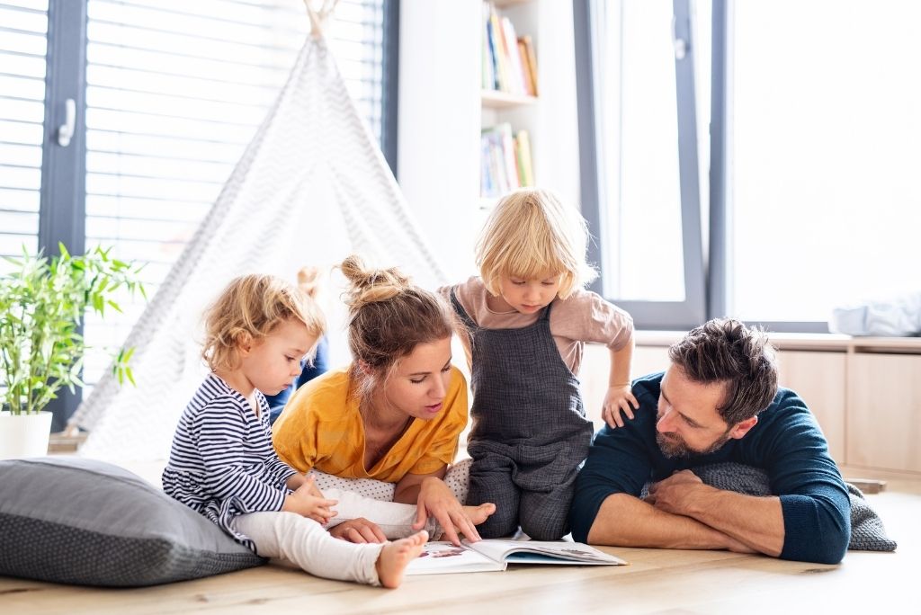 A family with two young children engrossed in reading a book together on the floor.