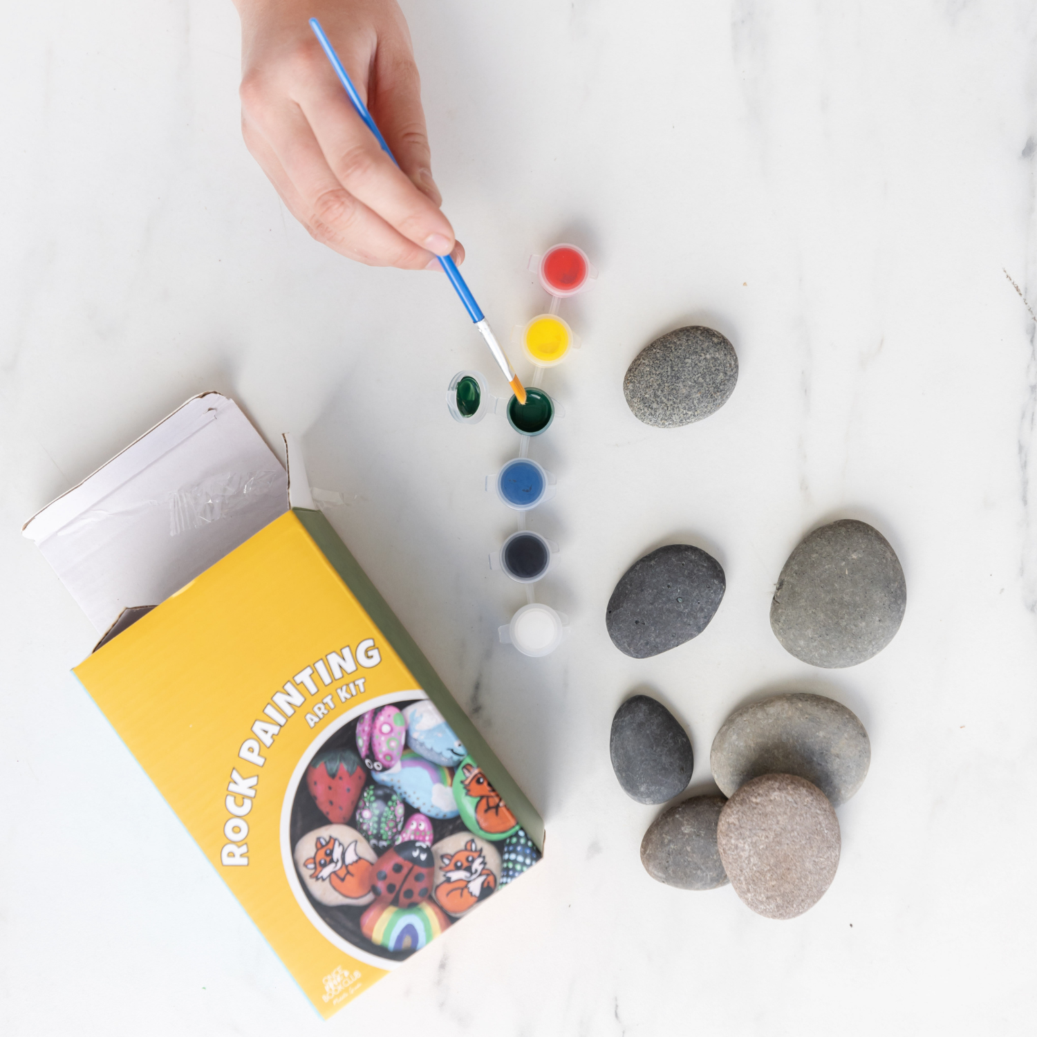 A white hand dips a paintbrush in paint. A box labeled Rock Painting Art Kit and a pile of rocks are next to the containers of paint.