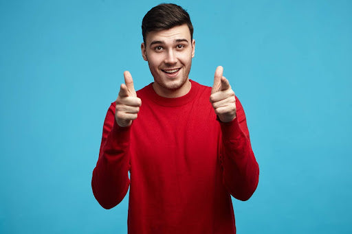 A cheerful man in a red sweater smiles broadly and points both thumbs towards the camera, giving a positive, encouraging gesture against a bright blue background.