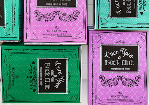 A close-up of vibrant teal and purple Once Upon a Book Club boxes with ornate designs, highlighting the club's motto "Bringing books to life, Monthly" and their website for a dynamic reading community.