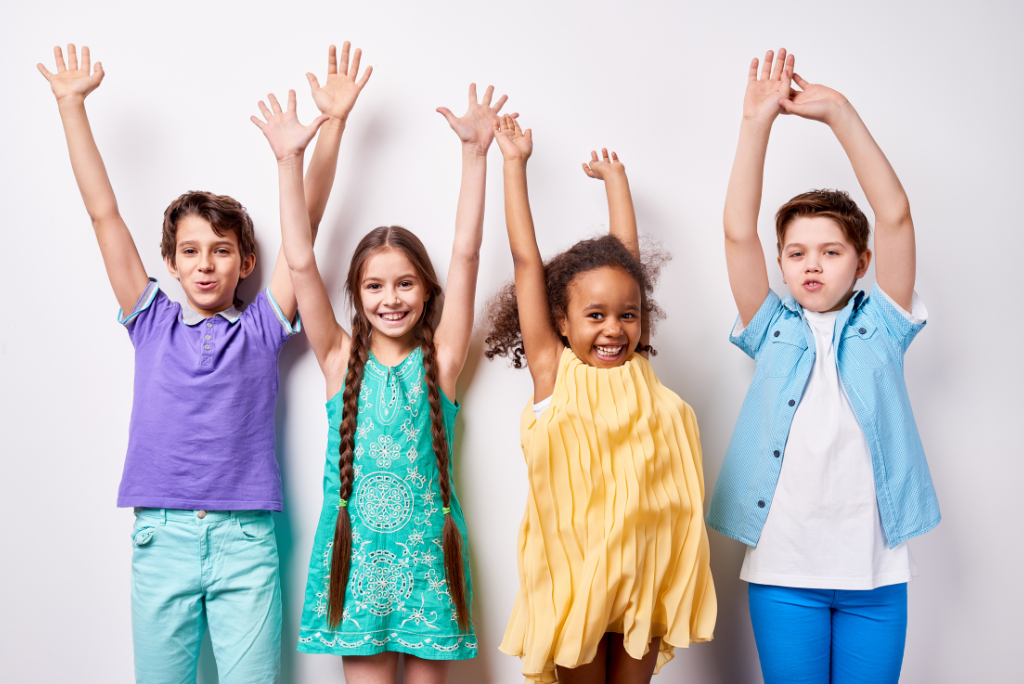 Four energetic kids standing in front of a white background with arms raised in celebration, dressed in colorful summer outfits.