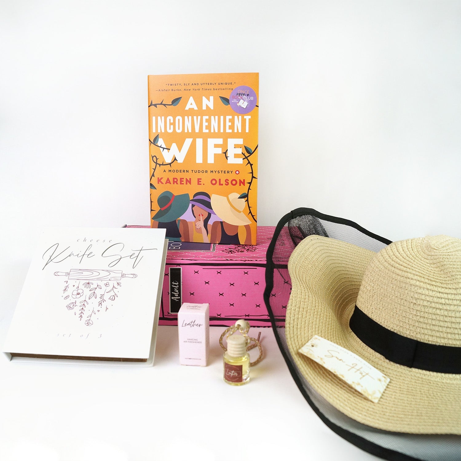 A hardcover edition of An Inconvenient Wife by Karen E Olson stands on a pink Once Upon a Book Club box. In front of the box are a white box labeled Knife Set, a box labeled car air freshener next to a small container, and a beige sunhat with black trim.