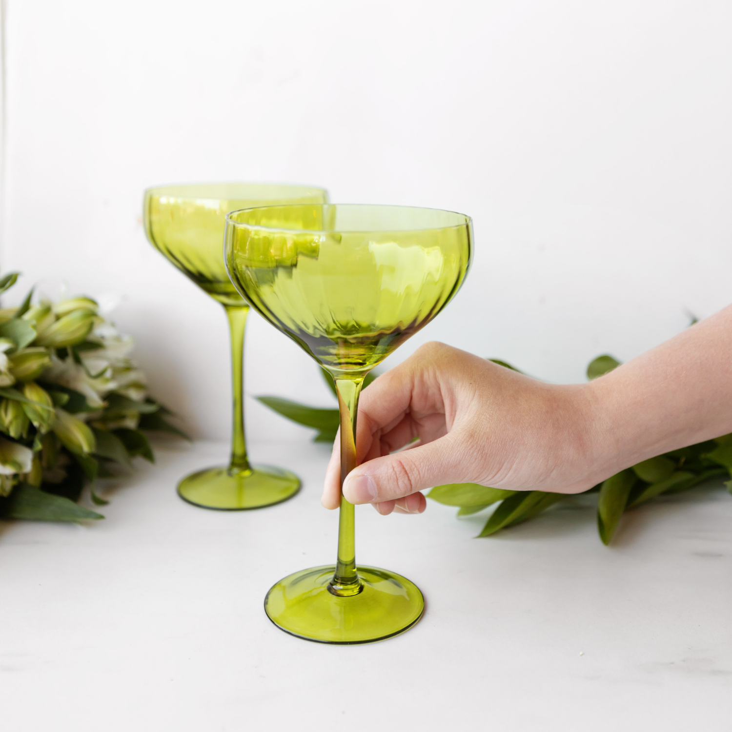a white hand holds the stem of a light green coupe glass with another matching glass behind it