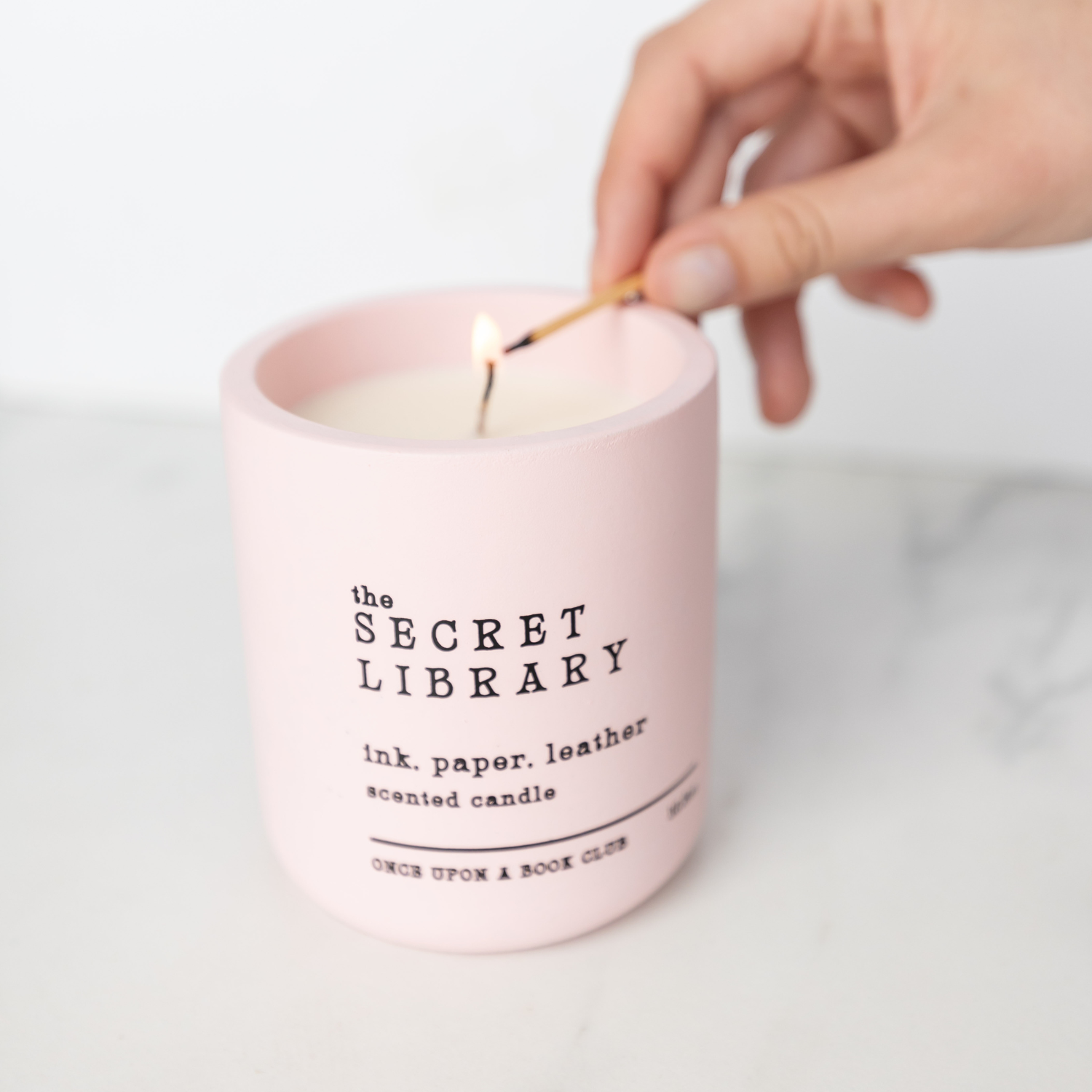 A white hand holding a match lights a candle. The candle is a pale pink with black text at the front that says The Secret Library, ink,paper,leather scented candle