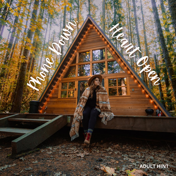 An A-frame cabin in the woods with a young brunette woman leaning on the front porch in a wide brimmed hat, plaid poncho, and boots. The words "Phones Down, Hearts Open" are written across the top of the image and the words "Adult Hint" are written along the bottom of the image.