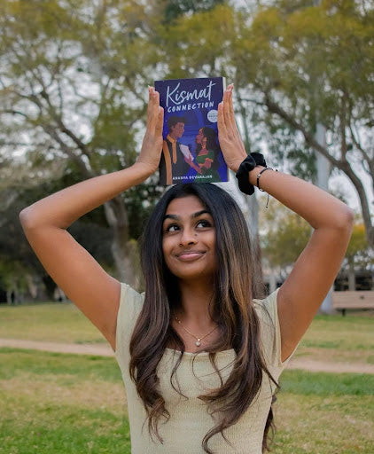 A smiling woman stands in a park, holding a book titled "Kismat Connection" by Ananya Devarajan above her head, aligning it with her face. The book cover depicts silhouettes of a couple against a colorful backdrop.