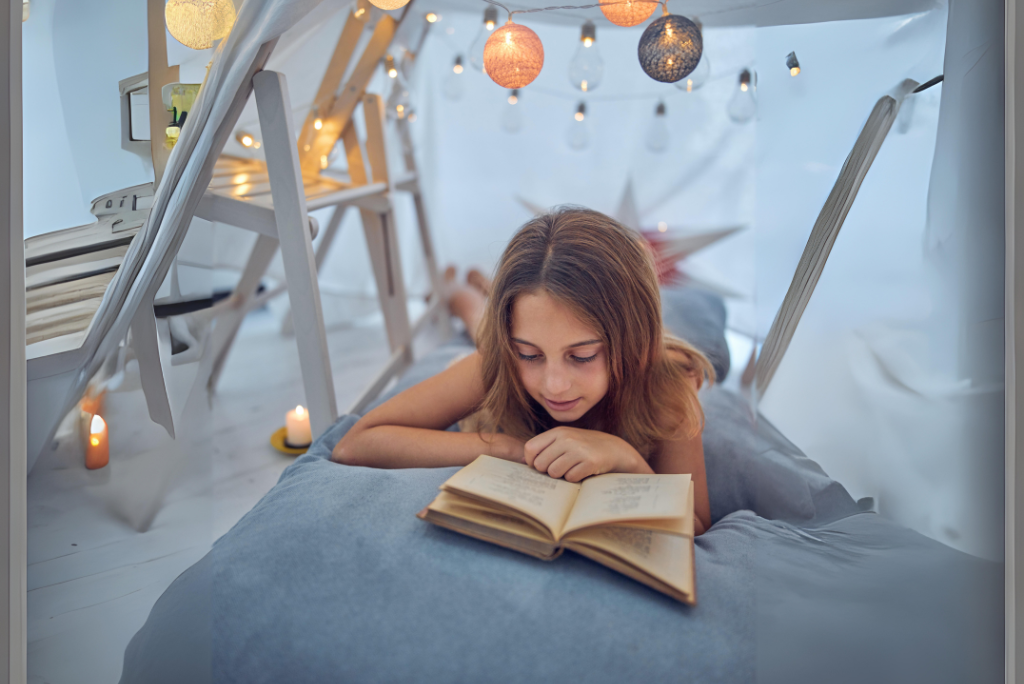 Young girl engrossed in reading under a cozy lit tent made of blankets.