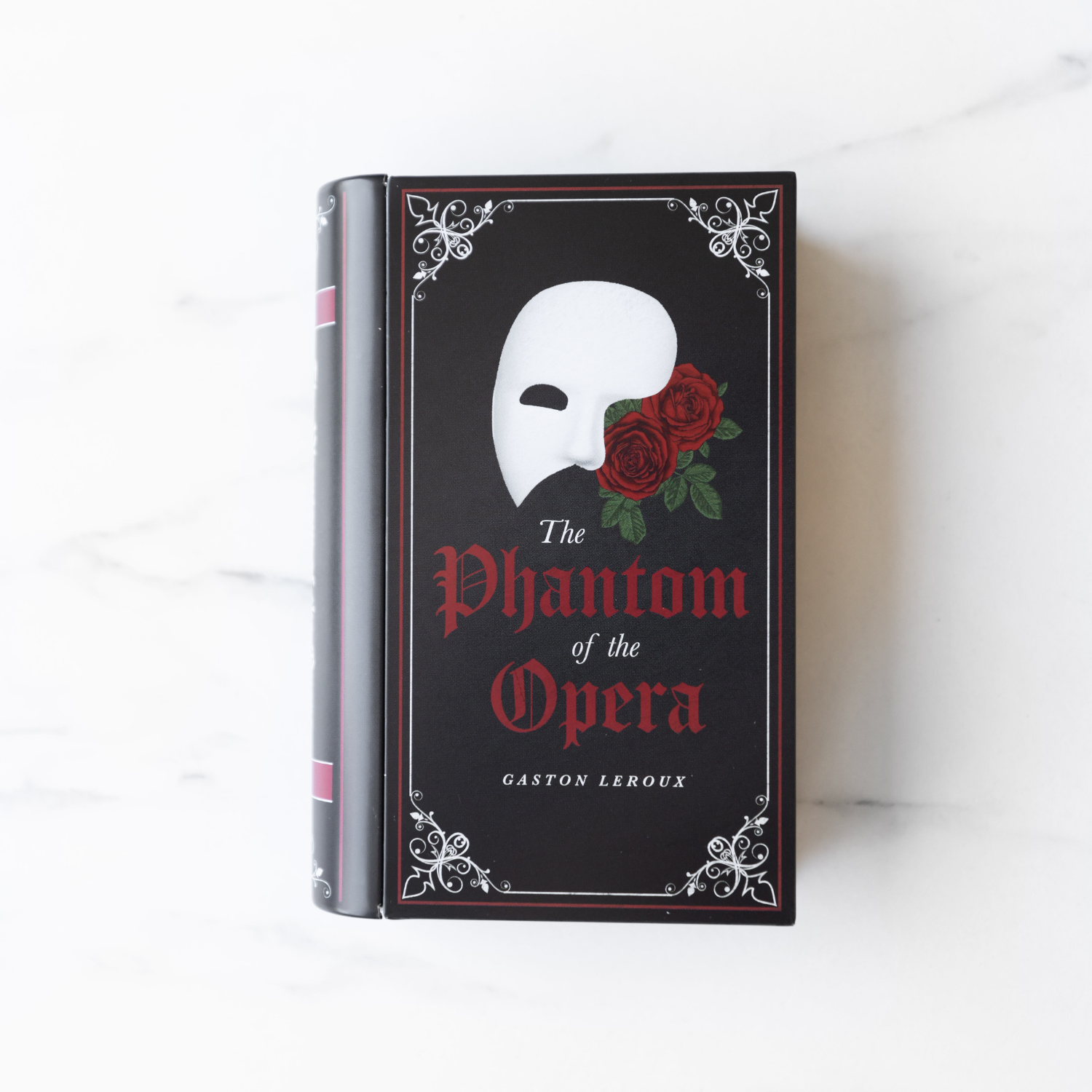 A black book tin that says The Phantom of the Opera on the front with a white mask and roses.