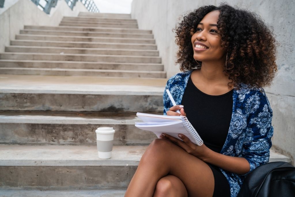 Smiling young woman sitting on steps writing in a notebook with a coffee cup beside her.