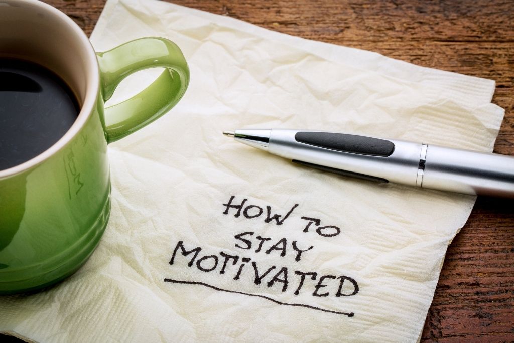 Coffee mug beside a napkin with "How to Stay Motivated" written on it and a pen, on a wooden table.