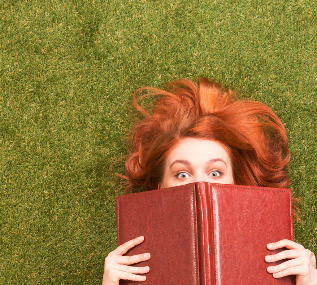 Red-haired woman lying on grass, wide-eyed over an open book.