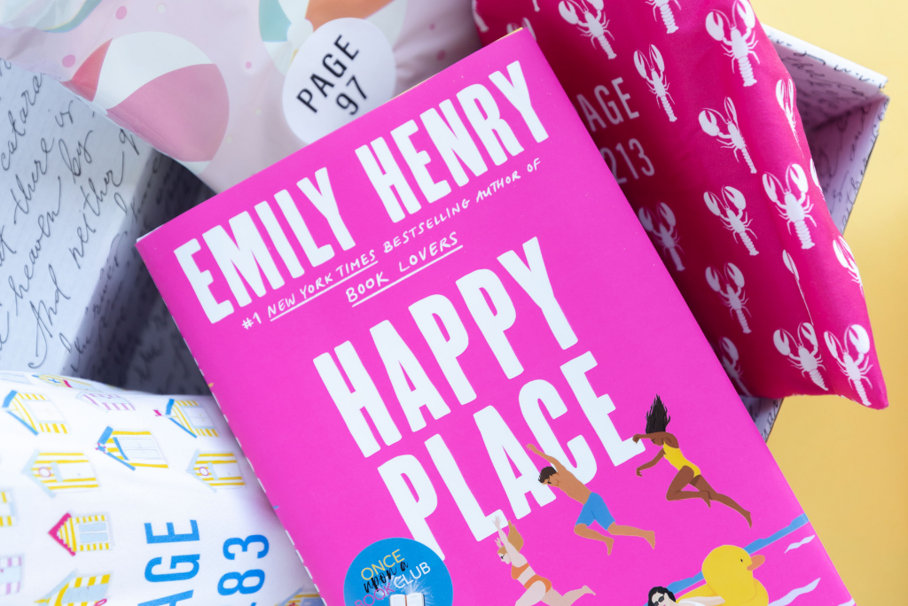 A pink book titled "Happy Place" by Emily Henry, with gift wrapping items.