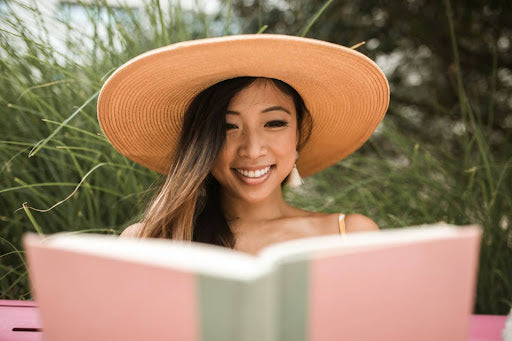 A smiling woman wearing a wide-brimmed hat reads a book outdoors, with the top edge of the pink book visible to the viewer.