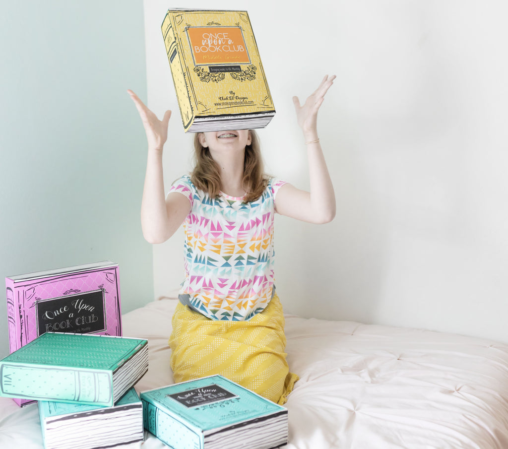 A young girl sits on a bed, happily tossing a "Once Upon a Book Club" box in the air with more colorful boxes around her, celebrating a fun and interactive reading experience.