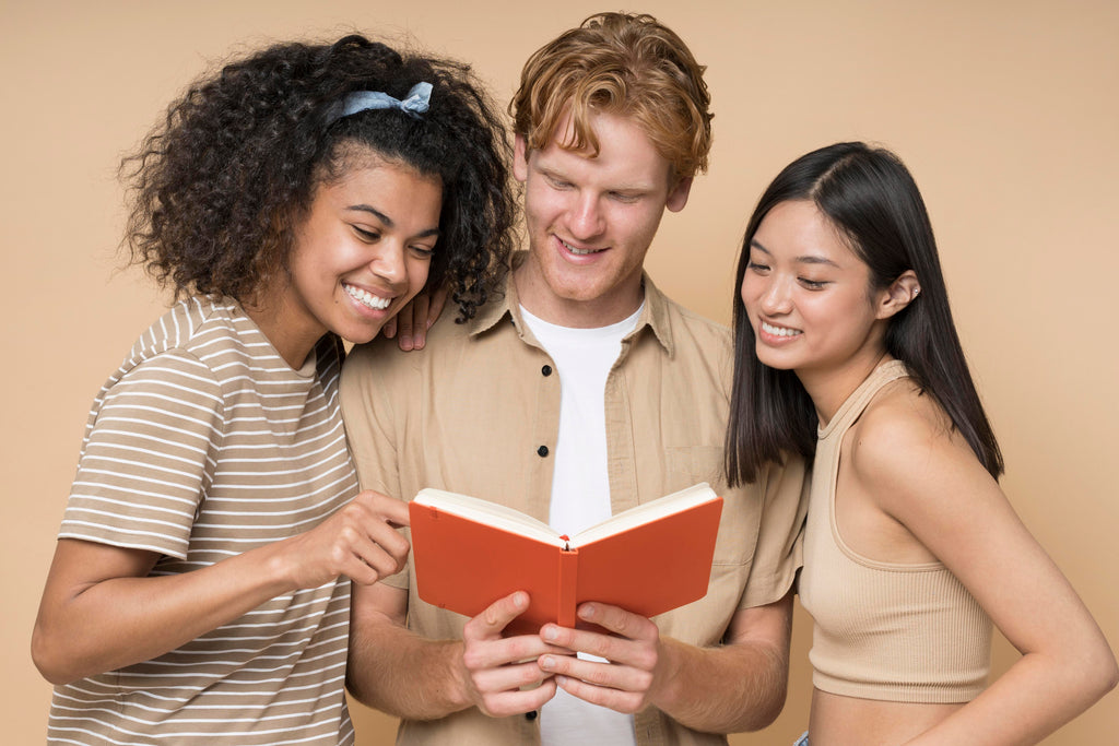 Three friends of diverse backgrounds share a joyful moment reading a book together, with expressions of amusement and engagement on their faces, against a neutral backdrop.
