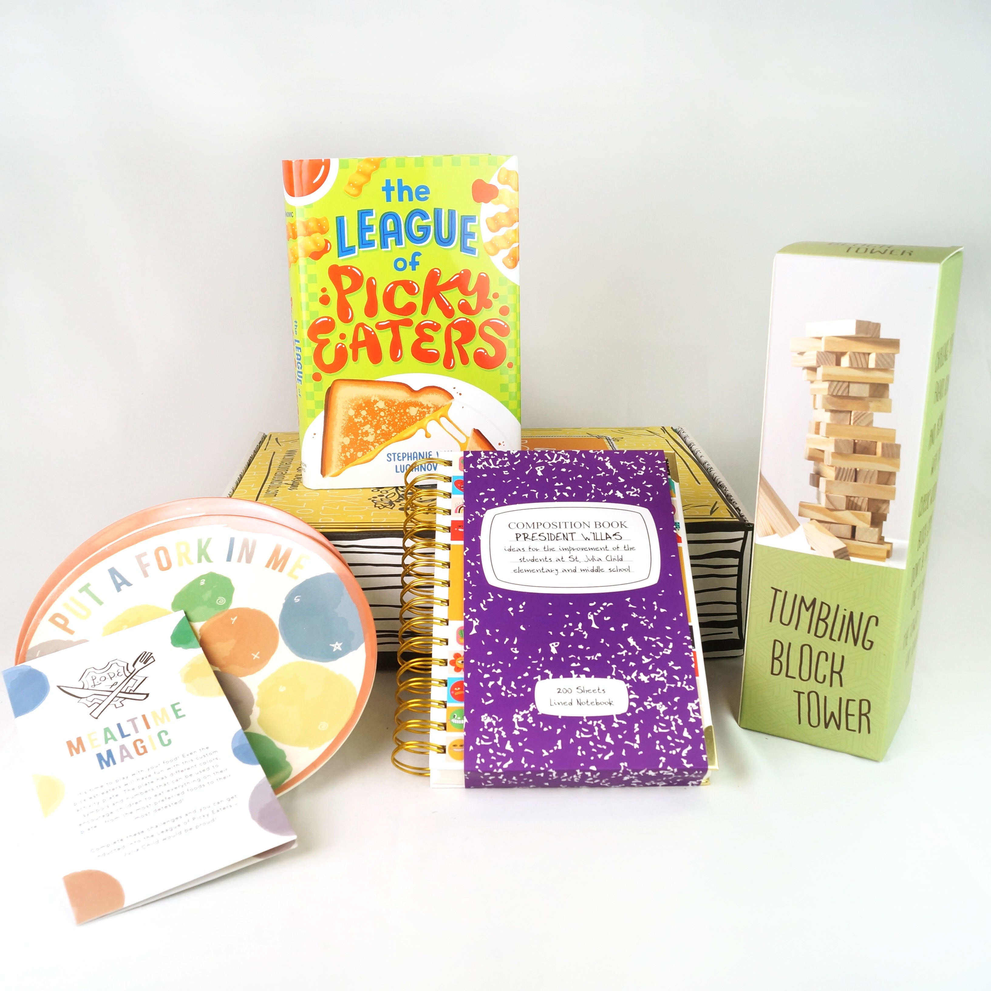 A hardcover edition of The League of Picky Eaters stands on a yellow OUABC box. In front of the box are two plates with colored dots on them, a purple notebook, and a box labeled tumbling block tower.