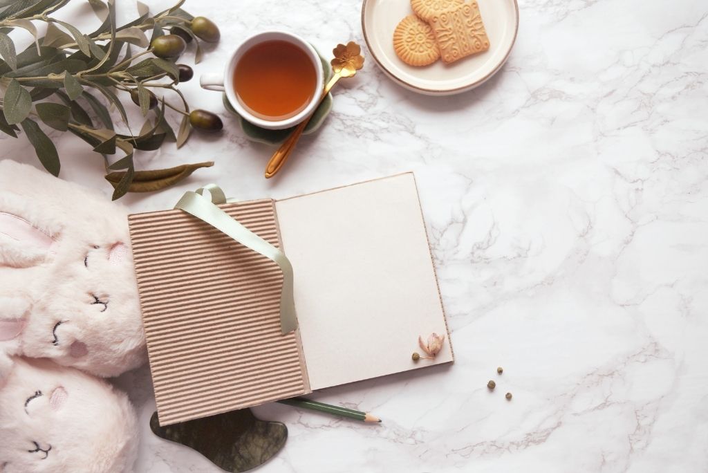 An open journal lies on a marble surface with a cup of tea, cookies, greenery, and cozy slippers, inviting a reflective moment.
