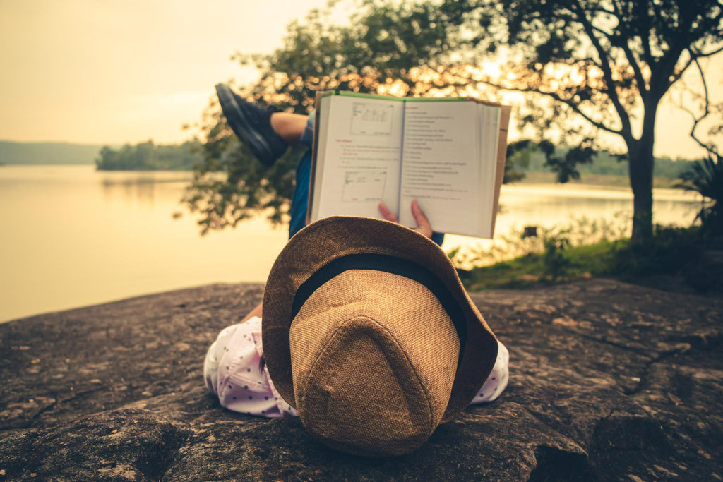 A person lying on a rock by a calm lake at sunset, immersed in reading a book with their hat off, evoking a sense of relaxation and escape in nature.