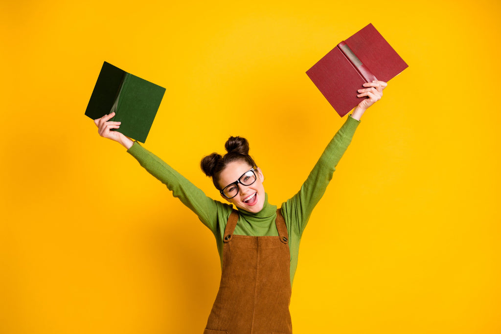 A young woman with glasses and a green turtleneck sweater, smiling and holding up two books, one green and one red, against a bright yellow background.